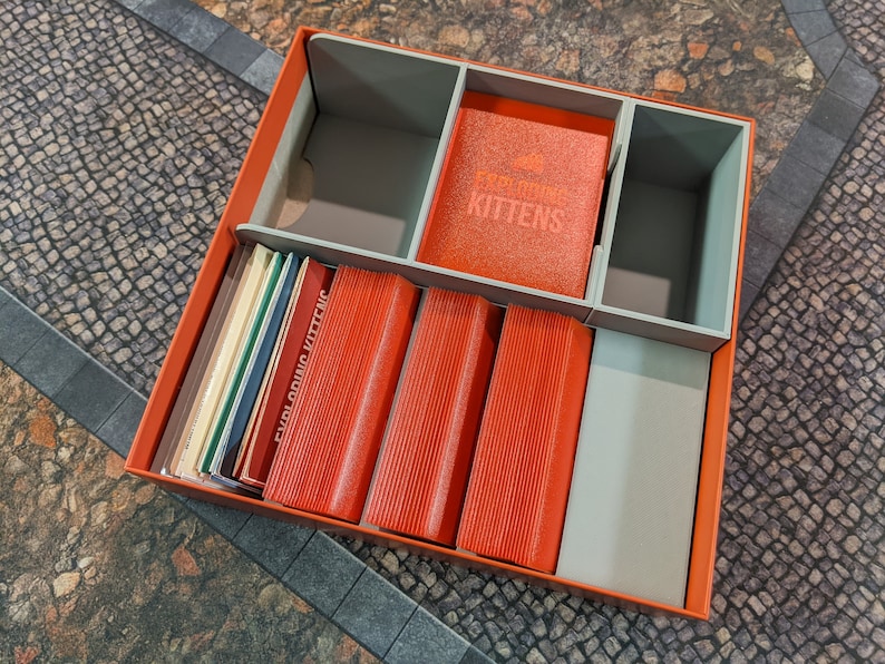 May include: An orange cardboard box with grey dividers for organising an Exploding Kittens card game. The box contains multiple decks of cards, including Exploding Kittens and a deck labelled "Exploding Kittens".