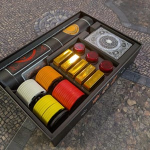 Peut inclure: Un jeu de société dans une boîte noire contenant un tube cylindrique noir avec des accents orange, un jeu de cartes aux motifs argentés complexes, quatre barres rectangulaires dorées, trois jetons circulaires rouges et des piles de jetons blancs, orange, rouges et jaunes.