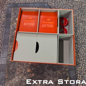 May include: A white and orange cardboard box with compartments for storing Exploding Kittens game cards and tokens. The box has the words "Exploding Kittens" printed on the front.