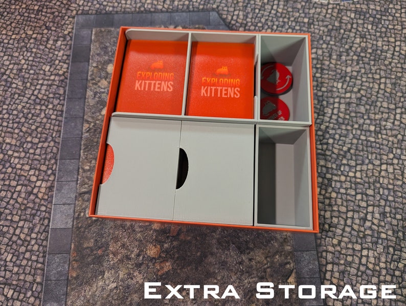 May include: A white and orange cardboard box with compartments for storing Exploding Kittens game cards and tokens. The box has the words "Exploding Kittens" printed on the front.