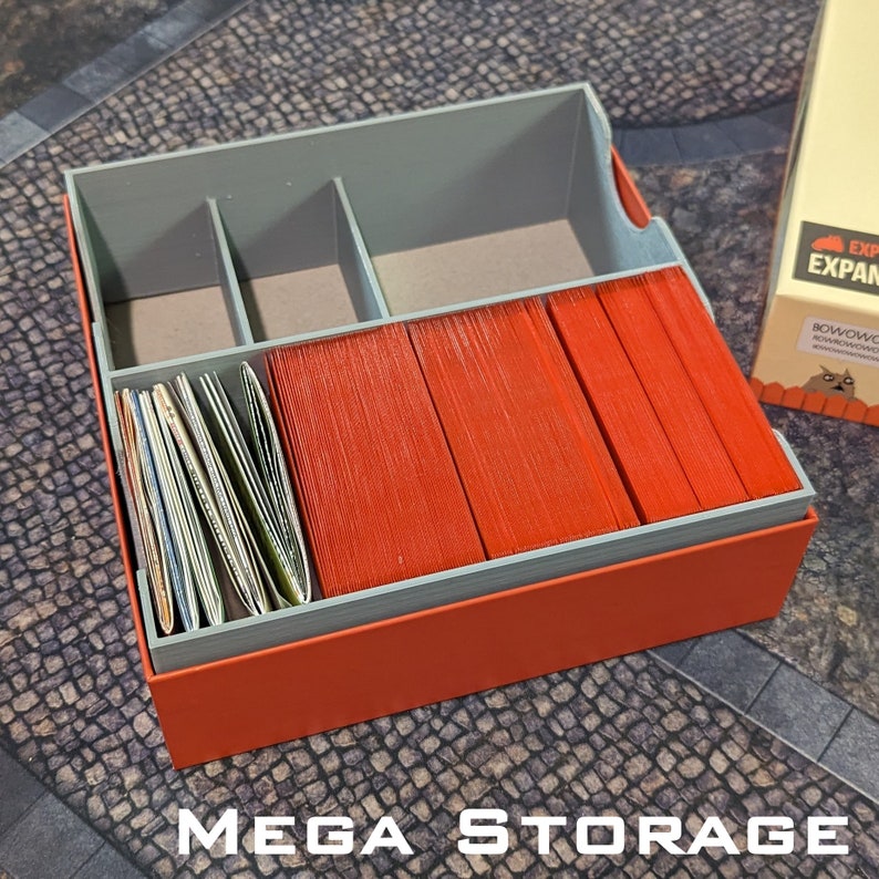 May include: A grey and orange storage box with a lid, designed to hold game cards. The box is filled with orange and white cards. The box has a label that says "Expansion".