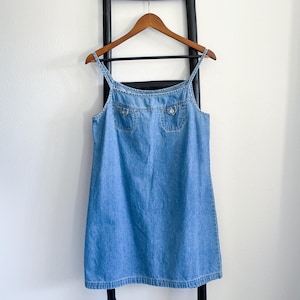 May include: Light blue denim dress with spaghetti straps and two front pockets. The dress is hanging on a wooden hanger against a white wall. The dress has a classic, casual style.