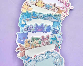 May include: A stack of colorful, die-cut stickers featuring various cartoon characters. Each sticker has a white space for writing, surrounded by a border of illustrated creatures in shades of blue, pink, yellow, and purple. The stickers are arranged on a light purple surface.