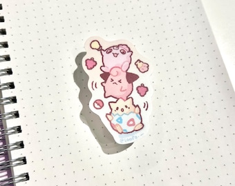 May include: A sticker featuring a stack of three cartoon characters in shades of pink, peach, and blue. The characters are arranged one on top of the other, with small hearts and other decorative elements around them. The sticker is on a white dotted paper.