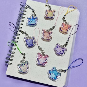 May include: A selection of colourful acrylic charms featuring cartoon animal designs. Each charm has a silver clasp and a matching coloured lanyard. The charms are displayed on a white dotted notebook against a purple background.