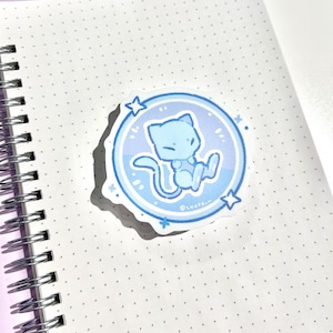 May include: A light blue sticker featuring a cartoon cat-like creature inside a blue circle with stars. The sticker is on a white notebook with a spiral binding. The creature has a long tail and is in a seated position.