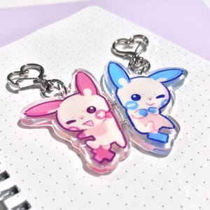 Op de afbeelding: Twee acryl sleutelhangers met cartoon konijn karakters. Eén konijn is roze en de andere blauw, beide met lachende gezichten en hartvormige sluitingen. De sleutelhangers liggen op een witte, gestippelde achtergrond.