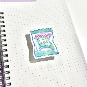 May include: A sticker of a candy bag with the text "Solosis" in a playful font. The bag is light blue and green, with a cartoon gummy character. The sticker is on a dotted notebook.