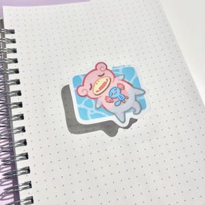 Puede incluir: Una pegatina con un personaje de dibujos animados en rosa, azul y blanco, flotando en un rectángulo azul. La pegatina está sobre un cuaderno blanco punteado. La pegatina tiene un borde blanco y una sombra.