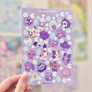 May include: A sheet of stickers with cartoon monster designs in purple, white, and pink. The stickers are on a lavender background with the text "SPOOKY-MONS" at the top. The artist's name, "@loora_," is at the bottom of the sheet.