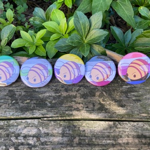 Op de afbeelding: Vijf ronde buttons met een cartoon visontwerp. Elke button heeft een ander kleurenschema, waaronder groen, blauw, geel, paars en roze. De vis heeft een gestreept patroon en een simpele, schattige uitdrukking. De buttons worden weergegeven op een houten oppervlak.