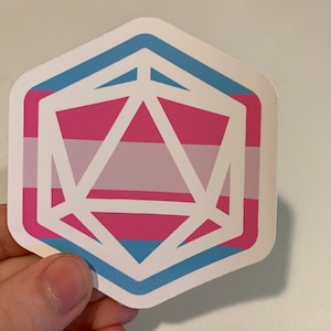 May include: A sticker featuring a white geometric design over a background of pink, light pink, and blue stripes, with a blue border. The sticker is held by a hand.