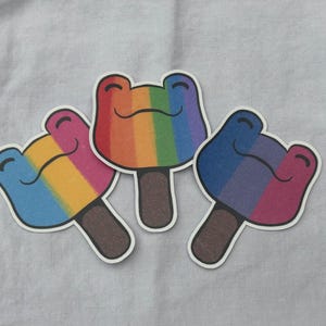 Puede incluir: Tres pegatinas coloridas con forma de paleta con caras de rana sonrientes. Cada pegatina presenta un esquema de color arcoíris diferente, que incluye un arcoíris clásico, un degradado rosa y amarillo y un degradado azul y morado. Las pegatinas tienen palitos de paleta marrones.
