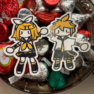 May include: Two cartoon stickers of anime-style characters with yellow hair and black and yellow outfits. The stickers are on a silver dish filled with foil-wrapped chocolates in red, silver, and green. Some chocolates are heart-shaped.