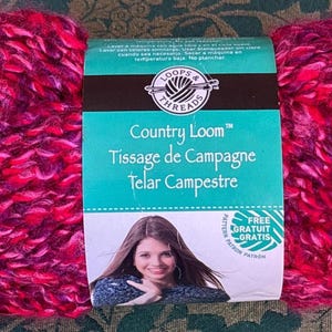 K&ouml;nnte beinhalten: Eine Kn&auml;uel Loops & Threads Country Loom Garn in einem rosa und violetten Farbton. Das Etikett lautet "Country Loom", "Tissage de Campagne" und "Telar Campestre".