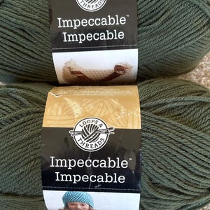 May include: Two skeins of Loops & Threads Impeccable yarn in a muted green color. Each skein has a label with the brand name and product name. The yarn is tightly wound and appears soft, suitable for various knitting and crochet projects.