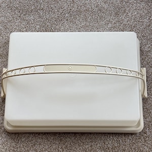 May include: A rectangular, off-white plastic cake carrier with a matching handle. The handle has a series of small, circular cutouts. The carrier is designed to hold and transport baked goods.