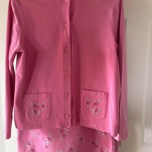 May include: A pink two-piece pajama set. The long-sleeved button-up top has two pockets with floral embroidery. The matching skirt features a floral pattern. The set is hanging on a white hanger.
