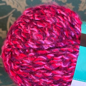 K&ouml;nnte beinhalten: Eine Kn&auml;uel pinkfarbenes, violettes und wei&szlig;es Garn. Das Garn ist weich und flauschig und perfekt zum Stricken oder H&auml;keln.