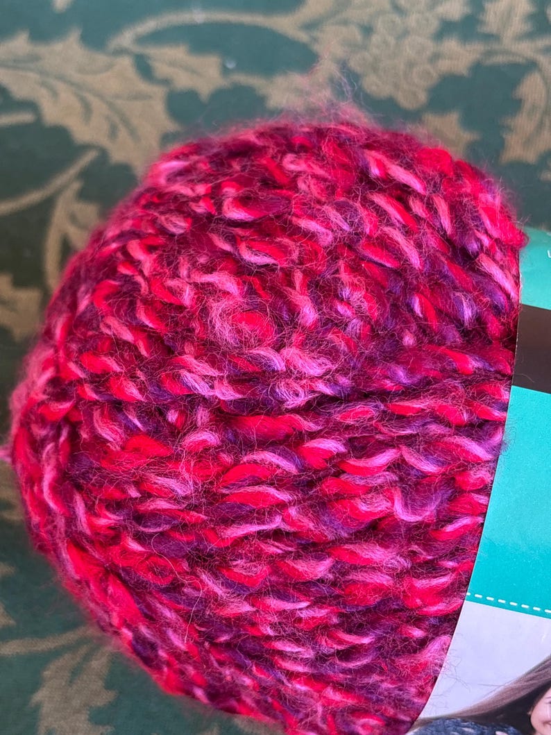 K&ouml;nnte beinhalten: Eine Kn&auml;uel pinkfarbenes, violettes und wei&szlig;es Garn. Das Garn ist weich und flauschig und perfekt zum Stricken oder H&auml;keln.