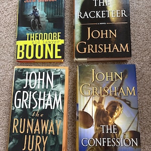 May include: Four hardcover books by John Grisham are displayed. Titles include "Theodore Boone: Kid Lawyer", "The Racketeeer", "The Runaway Jury", and "The Confession". The book covers feature illustrations and text in various colors.