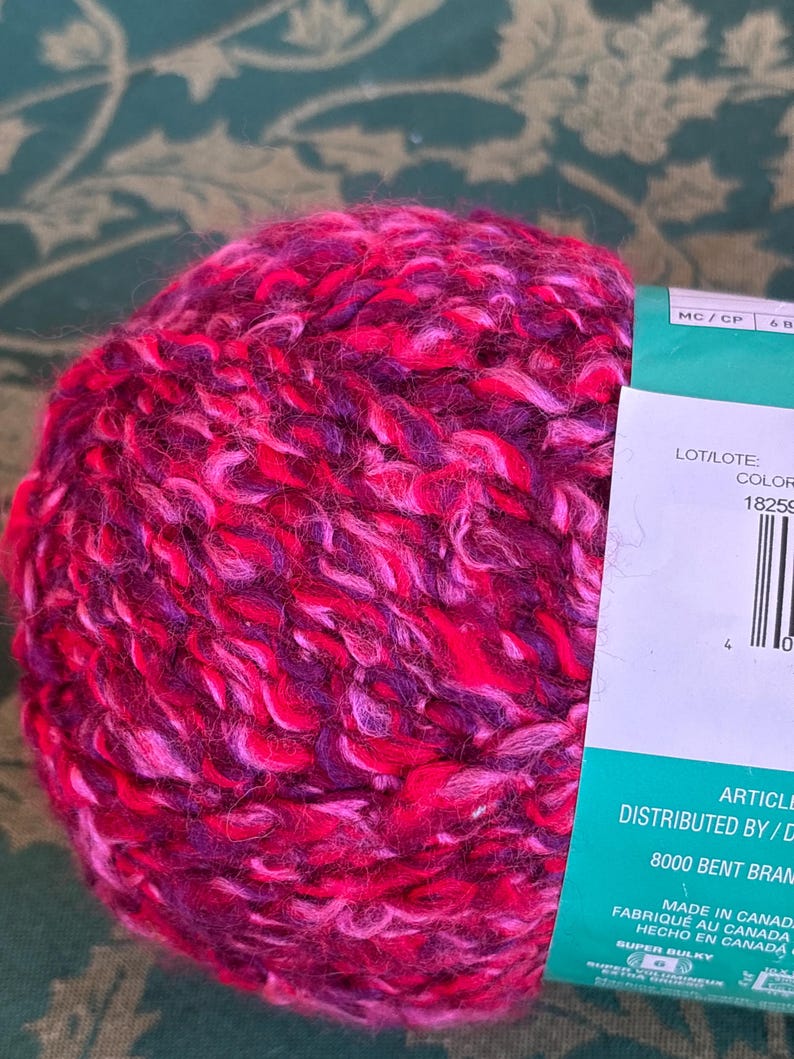 K&ouml;nnte beinhalten: Eine Kn&auml;uel pinkfarbenes, violettes und wei&szlig;es Garn mit einem Etikett, auf dem "LOT/LOTE: COLOR 18259" und "DISTRIBUTED BY/D 8000 BENT BRAM MADE IN CANADA FABRIQU&Eacute; AU CANADA HECHO EN CANADA SUPER BULKY" steht.