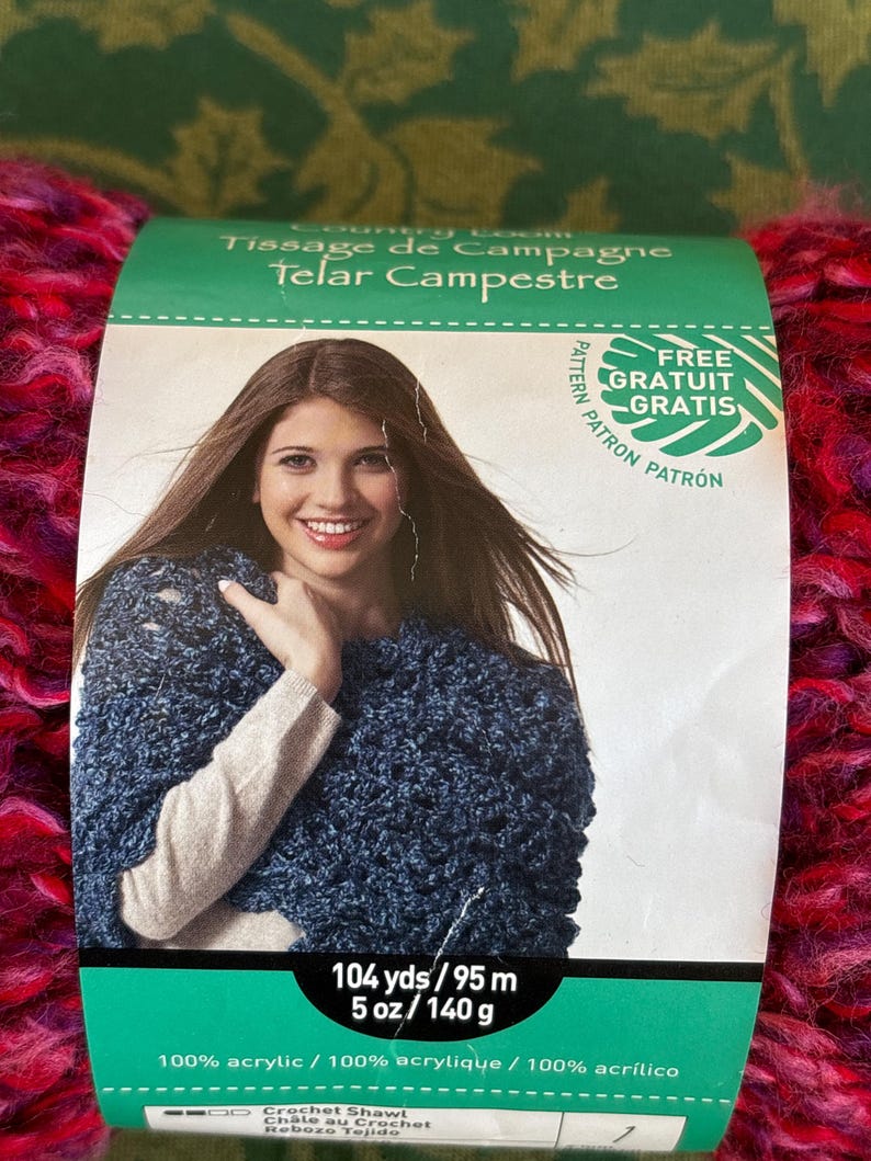 K&ouml;nnte beinhalten: Eine Kn&auml;uel pinkfarbenes und violettes Garn mit einem Etikett, auf dem "Tissage de Campagne Telar Campestre" steht und eine Frau mit einem blauen geh&auml;kelten Schal zu sehen ist. Das Etikett zeigt auch "Kostenloses Muster Gratuit Gratis" und enth&auml;lt das Garngewicht und die Faserzusammensetzung: 104 yds/95 m, 5 oz/140 g, 100 % Acryl.