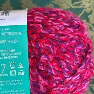 K&ouml;nnte beinhalten: Eine Kn&auml;uel pinkfarbenes, violettes und rotes Garn mit einem Etikett, auf dem "Imported Fiber, Fibres Import&eacute;es, As Importadas" steht und Pflegehinweise enthalten sind. Das Etikett enth&auml;lt auch den Text "# 211001 / Distribuido Por Irving, TX 75063".