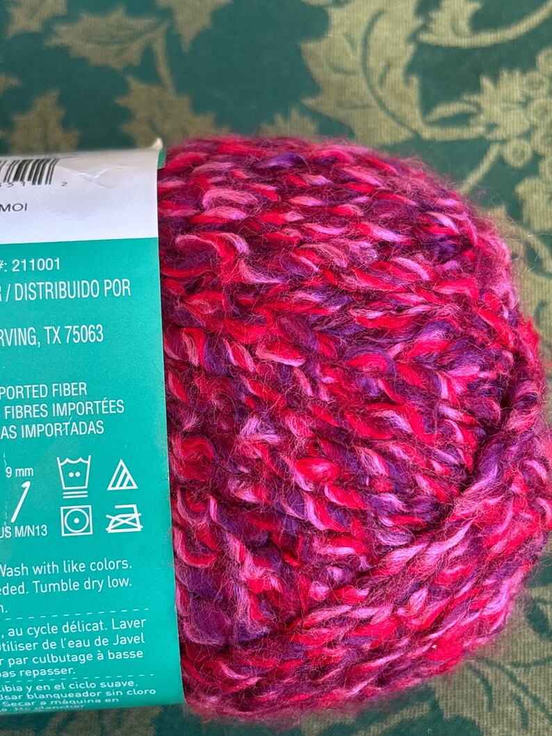 K&ouml;nnte beinhalten: Eine Kn&auml;uel pinkfarbenes, violettes und rotes Garn mit einem Etikett, auf dem "Imported Fiber, Fibres Import&eacute;es, As Importadas" steht und Pflegehinweise enthalten sind. Das Etikett enth&auml;lt auch den Text "# 211001 / Distribuido Por Irving, TX 75063".