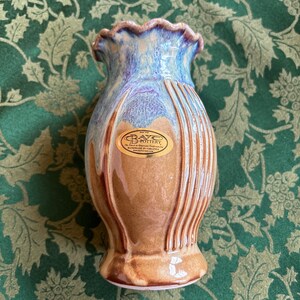 May include: A brown and blue ceramic vase with a ruffled top and a gold label that reads "The Heart of American Handcrafted Pottery, Handmade in Virginia, www.baycpottery.com".