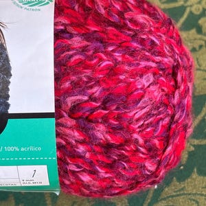K&ouml;nnte beinhalten: Eine rote, rosa und violette Garnrolle mit einem wei&szlig;en Etikett, auf dem "FREE GRATUIT GRATIS PATR&Oacute;N" und "100% acr&iacute;lico" steht. Das Etikett enth&auml;lt auch eine Nadelgr&ouml;&szlig;enempfehlung von 9 mm (US 13).