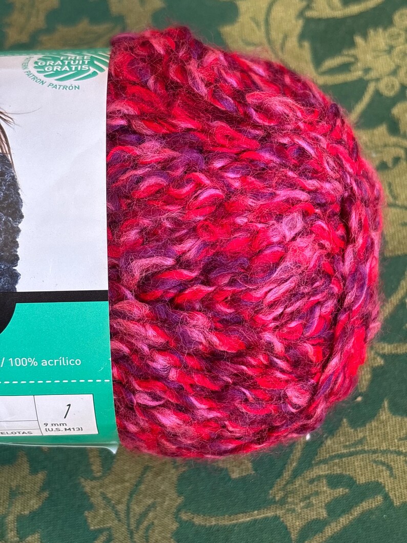 K&ouml;nnte beinhalten: Eine rote, rosa und violette Garnrolle mit einem wei&szlig;en Etikett, auf dem "FREE GRATUIT GRATIS PATR&Oacute;N" und "100% acr&iacute;lico" steht. Das Etikett enth&auml;lt auch eine Nadelgr&ouml;&szlig;enempfehlung von 9 mm (US 13).