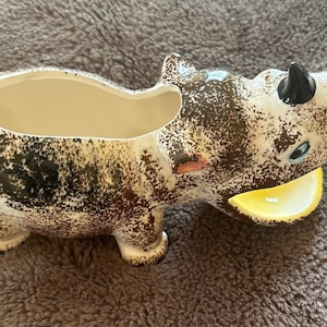 May include: A ceramic planter shaped like a rhinoceros with brown and white speckled paint. The planter has a yellow mouth and black horns.