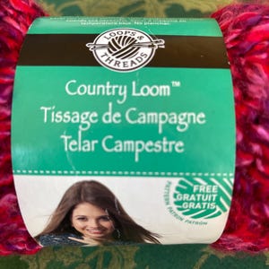 K&ouml;nnte beinhalten: Eine Garnrolle von Loops & Threads Country Loom in einem rosa-violetten Farbton. Das Etikett lautet "Country Loom", "Tissage de Campagne" und "Telar Campestre". Es steht auch "Kostenloses Muster/Gratuit/Gratis" drauf.