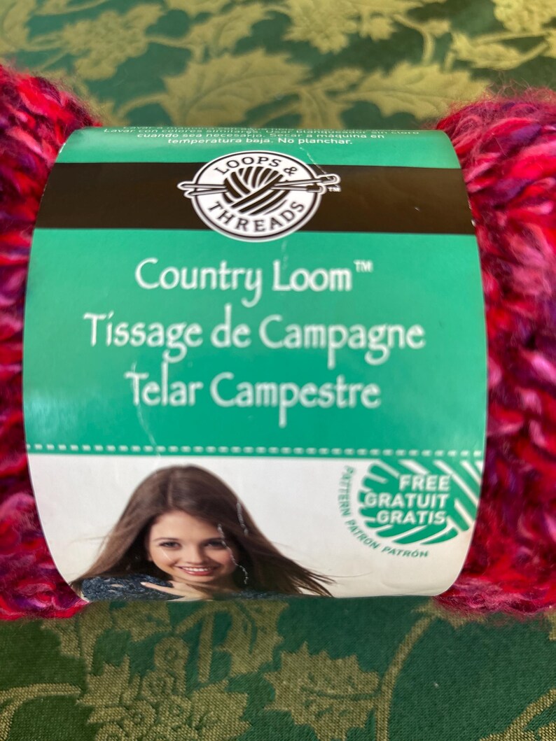 K&ouml;nnte beinhalten: Eine Garnrolle von Loops & Threads Country Loom in einem rosa-violetten Farbton. Das Etikett lautet "Country Loom", "Tissage de Campagne" und "Telar Campestre". Es steht auch "Kostenloses Muster/Gratuit/Gratis" drauf.