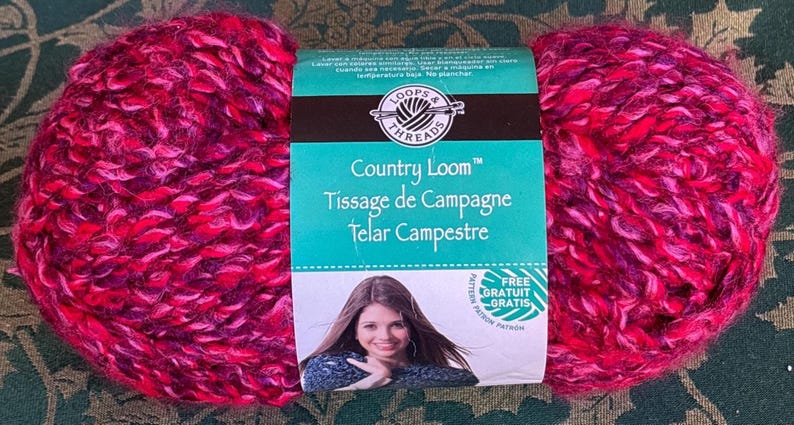 K&ouml;nnte beinhalten: Eine Kn&auml;uel Loops & Threads Country Loom Garn in einem rosa und violetten Farbton. Das Etikett lautet "Country Loom", "Tissage de Campagne" und "Telar Campestre".