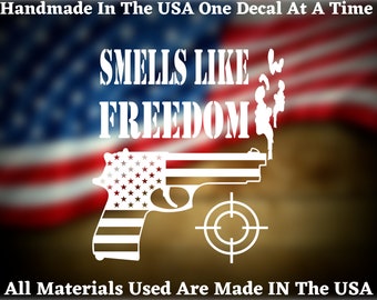 May include: White vinyl decal featuring the text "SMELLS LIKE FREEDOM" and a silhouette of a handgun with an American flag pattern. The decal is set against a blurred background of an American flag.