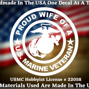 May include: White vinyl decal with the words "Proud Wife of a U.S. Marine Veteran" in a circle around the U.S. Marine Corps emblem. The decal is on a blurred background of the American flag.
