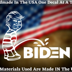 May include: White vinyl decal of a cartoon figure in the style of Donald Trump, wearing a red, white, and blue striped outfit, urinating on the word "BIDEN". The background is a blurred image of the American flag.