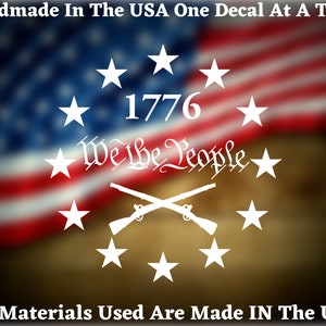 Peut inclure: Autocollant en vinyle blanc avec un cercle d'étoiles entourant un motif de fusils croisés. Le texte "1776 We The People" est à l'intérieur du cercle d'étoiles. L'autocollant est sur un fond flou d'un drapeau américain.