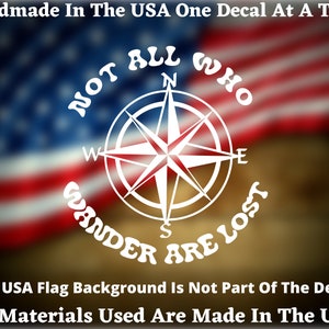 May include: White vinyl decal with a compass and the text "NOT ALL WHO WANDER ARE LOST". The decal is on a blurred background of an American flag.