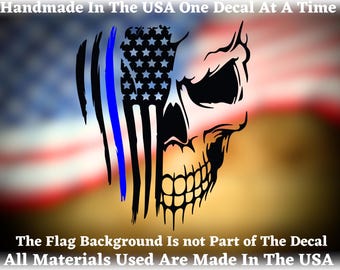 May include: A black and white skull decal with a thin blue line and an American flag design. The decal is on a blurred background of an American flag. The text "Handmade in the USA One Decal at a Time" and "The Flag Background Is not Part of The Decal All Materials Used Are Made In The USA" is at the top and bottom of the image.