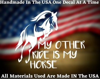 May include: White vinyl decal featuring a silhouette of a horse jumping, with the text "MY OTHER RIDE IS MY HORSE" underneath. The decal is displayed against a blurred American flag background.