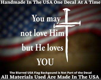 May include: White vinyl decal with a cross made of nails and the text "You may not love Him but He loves YOU". The decal is on a blurred background of an American flag.