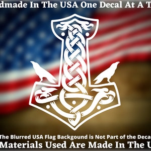 May include: White vinyl decal of a Mjolnir hammer with a Celtic knot design and two ravens. The decal is on a blurred background of the American flag. The text "Handmade in the USA One Decal at a Time" and "All Materials Used Are Made In The USA" are at the top and bottom of the image.