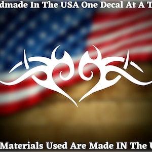 May include: White vinyl decal of a stylized heart with tribal-inspired designs. The decal is set against a blurred background of the American flag. The text "Handmade In The USA One Decal At A Time" appears at the top of the image. The text "All Materials Used Are Made IN The USA" appears at the bottom of the image.