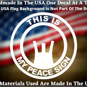 May include: White vinyl decal with the text "THIS IS MY PEACE SIGN" in a circle. The decal features a stylized peace sign with a hand-like shape. The decal is shown against a blurred American flag background.