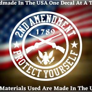 May include: White vinyl decal with a circle design featuring crossed rifles, an eagle, and the text "2ND AMENDMENT 1789 PROTECT YOURSELF". The decal is on a blurred background of the American flag.
