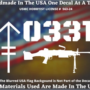 May include: White decal featuring the number "0331", a stylized arrow, and a machine gun silhouette. The text "Handmade In The USA" and "USMC HOBBYIST LICENSE # 563-24" are also present. The background is a blurred American flag.