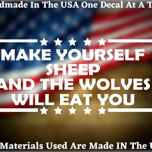 May include: White vinyl decal with the text "MAKE YOURSELF SHEEP AND THE WOLVES WILL EAT YOU" surrounded by stars. The decal is on a blurred background of an American flag.